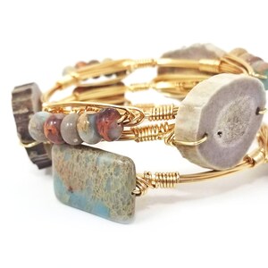 May include: A set of three gold wire wrapped bracelets featuring natural elements. The bracelets feature a light brown antler slice, a light blue and brown agate slice, and a mix of brown, light blue, and white beads.