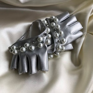 May include: A pair of silver fingerless gloves with a pearl chain detail. The gloves are laying on a white satin fabric.