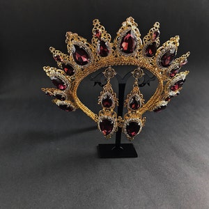 May include: A gold crown with a burgundy red gemstone design and matching earrings. The crown is ornate and features a delicate filigree pattern.