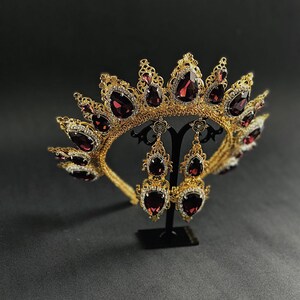 May include: A gold crown with a filigree design and dark red teardrop-shaped gemstones. The crown is displayed on a black stand with matching earrings.