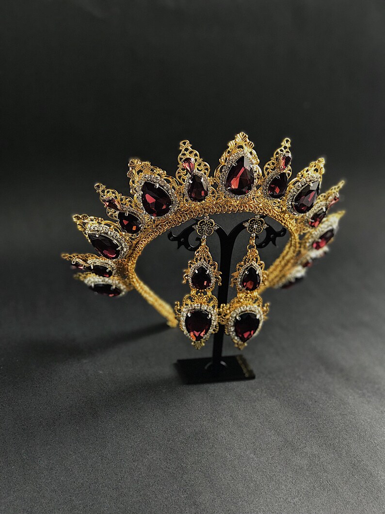 May include: A gold crown with a filigree design and dark red teardrop-shaped gemstones. The crown is displayed on a black stand with matching earrings.