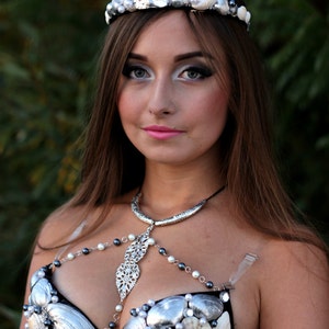 May include: A woman wearing a silver and white seashell bra with a matching crown. The bra is made of seashells and pearls and is attached to a silver chain.