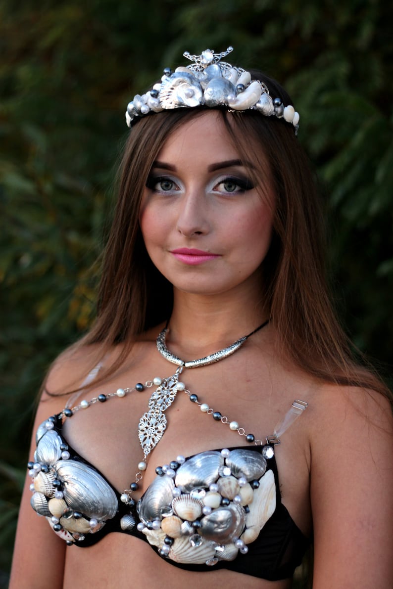 May include: A woman wearing a silver and white seashell bra with a matching crown. The bra is made of seashells and pearls and is attached to a silver chain.
