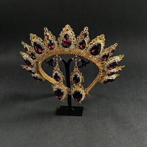 May include: A gold crown with a burgundy red gemstone design and matching earrings. The crown and earrings are on a black stand.