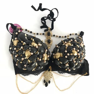 May include: A black bra decorated with seashells, gold accents, and pearls. The bra has a black lace trim and gold chains with a pendant.