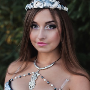 May include: A woman wearing a silver and white seashell crown and a black bra decorated with silver and white seashells. The bra has a silver chain and pendant necklace.