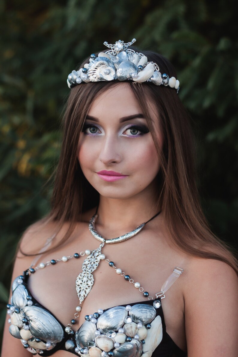 May include: A woman wearing a silver and white seashell crown and a black bra decorated with silver and white seashells. The bra has a silver chain and pendant necklace.