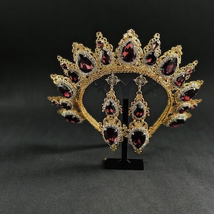 May include: A gold crown with a burgundy jeweled design and matching earrings. The crown is on a black stand.
