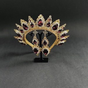 May include: A gold crown with a burgundy red gemstone design and matching earrings. The crown and earrings are on a black stand.