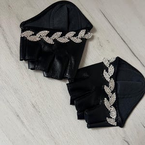 May include: A pair of black fingerless gloves featuring a smooth finish and a decorative band of clear, sparkling rhinestones arranged in a leaf-like pattern across the back of the hand. The gloves have open fingertips and a structured wrist area.
