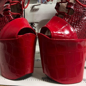 May include: A pair of bright red platform shoes with a glossy crocodile-embossed texture, featuring an open-toe design and red lace-up closures that extend up the front. The shoes are positioned on a surface with a visible ruler showing measurements.