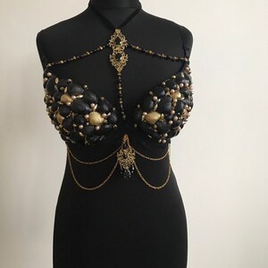 May include: A black bra decorated with gold and black seashells, beads, and chains. The bra has a delicate gold pendant with black stones hanging from the center.