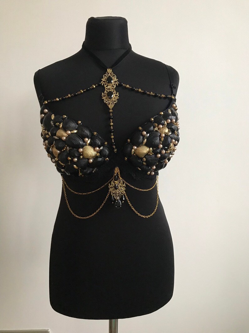 May include: A black bra decorated with gold and black seashells, beads, and chains. The bra has a delicate gold pendant with black stones hanging from the center.