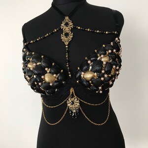 May include: A black bra decorated with seashells and gold chains. The bra has a black lace overlay and a gold pendant with black beads hanging from the center.