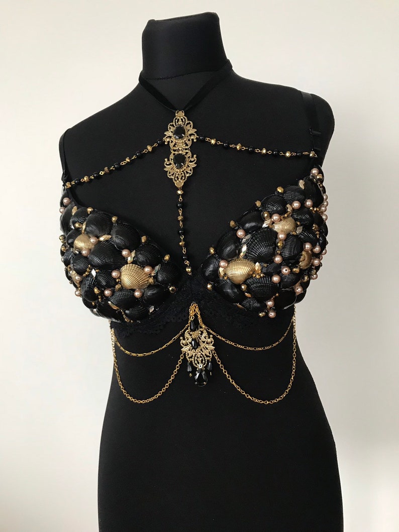 May include: A black bra decorated with seashells and gold chains. The bra has a black lace overlay and a gold pendant with black beads hanging from the center.