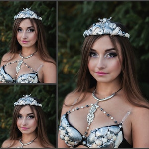May include: A woman wearing a silver and white seashell crown and a silver and white seashell bra. The bra has a silver chain with a pendant in the shape of a seashell. The woman is wearing a black top and has long brown hair.