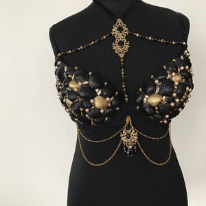 May include: A black bra decorated with seashells, pearls, and gold chains. The bra has a black lace trim and a gold pendant with a black gemstone.