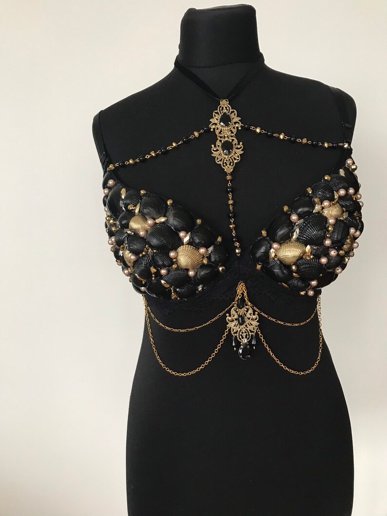 May include: A black bra decorated with seashells, pearls, and gold chains. The bra has a black lace trim and a gold pendant with a black gemstone.