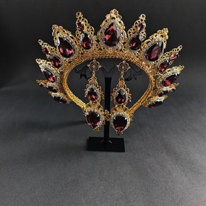 May include: A gold crown with a burgundy jeweled design and matching earrings. The crown is ornate and features a delicate filigree pattern. The earrings are teardrop shaped and dangle from gold hooks.