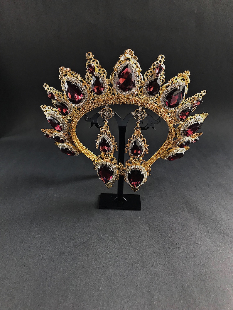 May include: A gold crown with a burgundy jeweled design and matching earrings. The crown is ornate and features a delicate filigree pattern. The earrings are teardrop shaped and dangle from gold hooks.