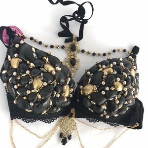 May include: A black bra decorated with seashells, pearls, and gold accents. The bra has a black lace trim and gold chains with a decorative pendant.