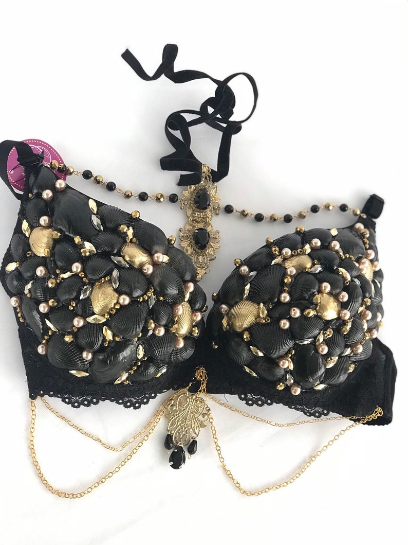 May include: A black bra decorated with seashells, pearls, and gold accents. The bra has a black lace trim and gold chains with a decorative pendant.