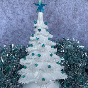 Könnte beinhalten: Ein weißer Keramik-Weihnachtsbaum mit blauen, schillernden Glühbirnen und einem blauen Stern oben. Der Baum ist von silbernem und blauem Lametta umgeben.