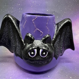 Puede incluir: Un recipiente de cerámica morado con forma de murciélago con alas negras brillantes y una cara triste. El recipiente tiene una abertura cilíndrica y está decorado con detalles brillantes. La cara del murciélago es negra con ojos blancos y boca hacia abajo.