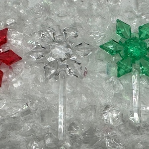 May include: Three plastic snowflake ornaments with a red, clear, and green color scheme. The snowflakes are on a white background.