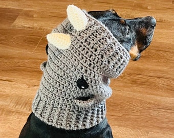 May include: A black dog wearing a gray crocheted dinosaur costume with white horns. The costume has a black eye patch.