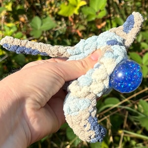 Puede incluir: Un juguete de crochet azul y blanco con una pelota de plástico azul llena de bolitas azules más pequeñas. El juguete está siendo sostenido por una mano.