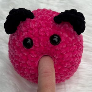 May include: A handmade, plush, pink toy with black accents. The toy has two black, rounded ears and two black, round eyes. A finger is inserted into a hole in the toy's center.