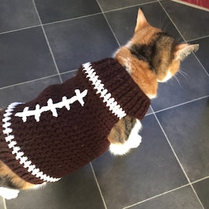 May include: A brown and white crocheted cat sweater with a football design. The sweater has white stitching that resembles the laces of a football.