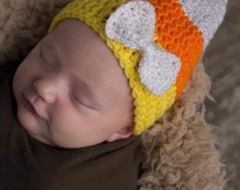 May include: A baby wearing a crocheted candy corn hat with a white bow. The hat is yellow, orange, and white.