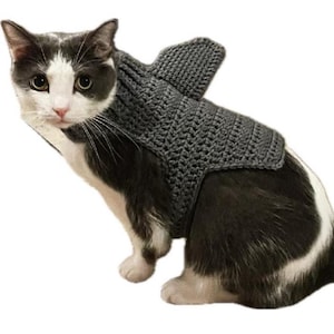 May include: A black and white cat wearing a gray crocheted shark costume. The costume has a fin on the back and covers the cat's torso.
