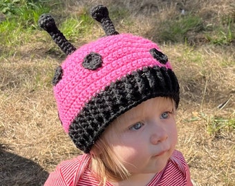 May include: A pink and black crocheted ladybug hat with black antennae. The hat is worn by a child with blonde hair.