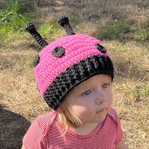 Può includere: Un cappello di coccinella in crochet rosa e nero con antenne nere. Il cappello è indossato da un bambino con i capelli biondi.