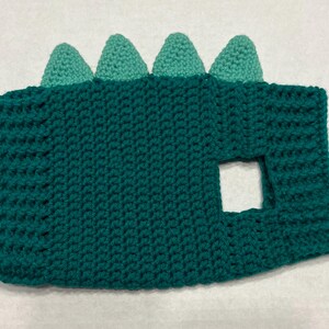 Pu&ograve; includere: Costume da dinosauro in crochet verde con un foro per la testa e una schiena a punta verde e bianca.