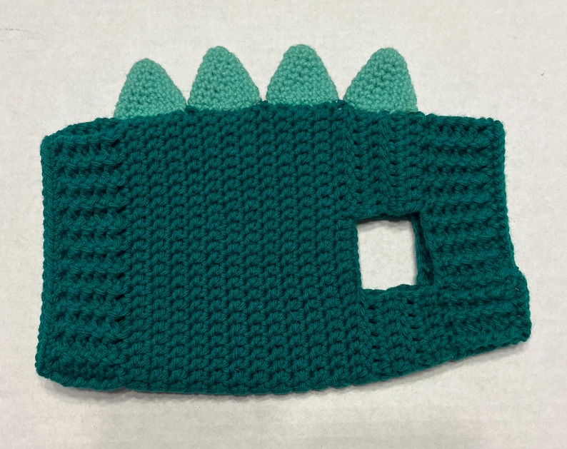 Pu&ograve; includere: Costume da dinosauro in crochet verde con un foro per la testa e una schiena a punta verde e bianca.