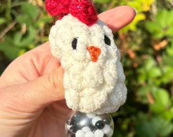 May include: White crocheted chicken toy with a red bow on its head. The chicken is holding a clear plastic ball filled with black and white beads.
