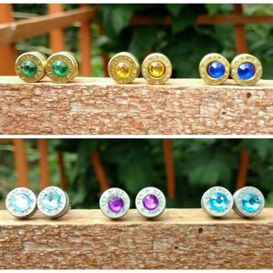 May include: A collection of stud earrings with various colored gemstones. The earrings are made from brass casings and feature green, yellow, blue, light blue, purple, and clear gemstones. The earrings are displayed on a wooden surface.