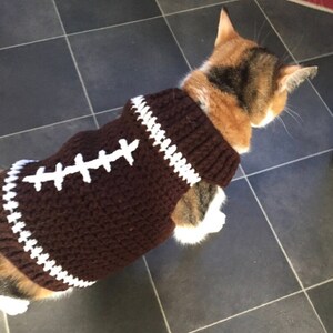 Pu&ograve; includere: Un maglione da gatto in crochet marrone con cuciture bianche, che ricorda un pallone da calcio. Il maglione ha un bordo bianco intorno al collo e ai bordi.