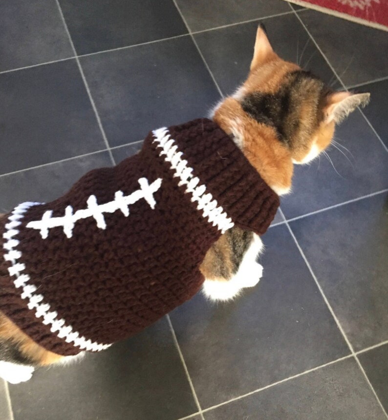 Pu&ograve; includere: Un maglione da gatto in crochet marrone con cuciture bianche, che ricorda un pallone da calcio. Il maglione ha un bordo bianco intorno al collo e ai bordi.