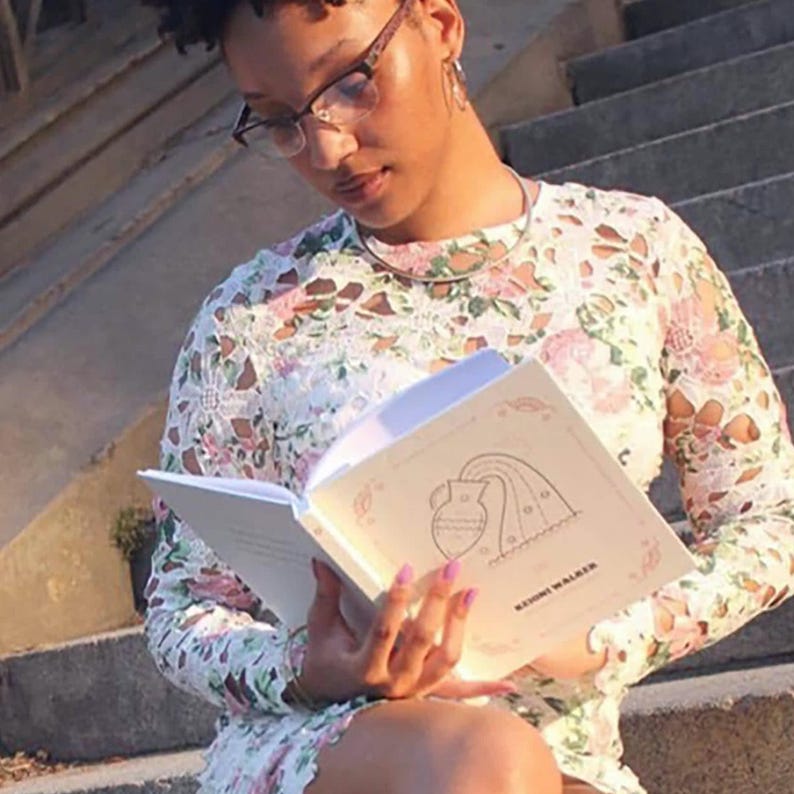 May include: A person reading a white journal with a pink and white floral design. The journal has a rainbow and a pitcher with water pouring out of it. The text on the journal reads "Rising Water".