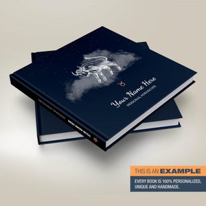 May include: A dark blue hardback book with a white illustration of a bull and stars on the cover. The text "Your Name Here" and "Personal Horoscope" is printed on the cover. The book is stacked on top of another book.