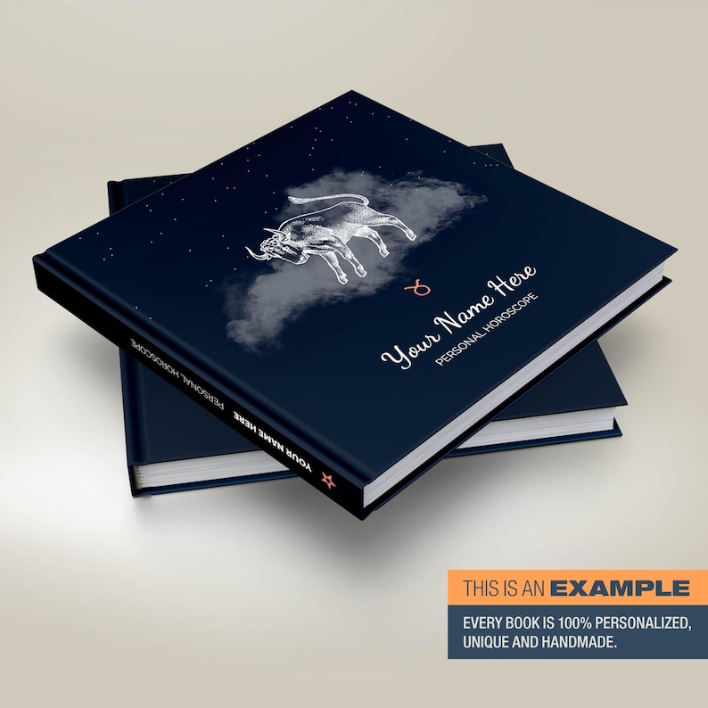 May include: A dark blue hardback book with a white illustration of a bull and stars on the cover. The text "Your Name Here" and "Personal Horoscope" is printed on the cover. The book is stacked on top of another book.