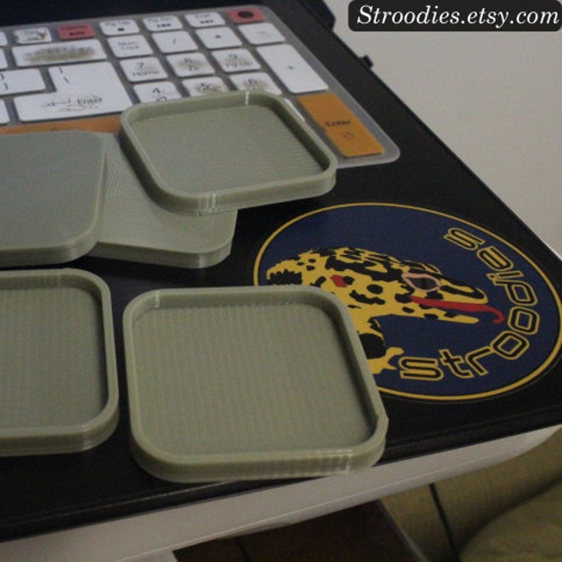 May include: Four gray square coasters made of 3D printed plastic. The coasters are on a black surface with a sticker that says "Stroodies.etsy.com".