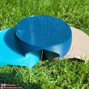 May include: A teal-colored, 3D-printed object with a textured surface, resting on a light blue and tan surface. The object has a unique, curved design. The image is taken outdoors on a bed of green grass.