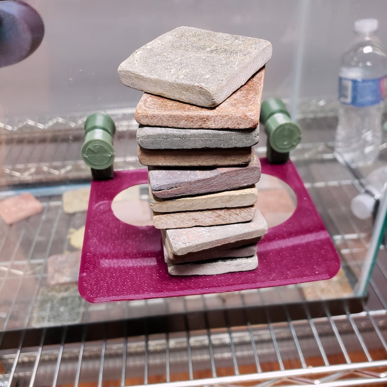 May include: A stack of ten square tiles in various shades of brown, beige, and gray. The tiles are stacked on a burgundy circular platform.