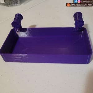 May include: A purple plastic tray with two small, round, cylindrical handles on either side. The tray is empty and appears to be made for holding small items.